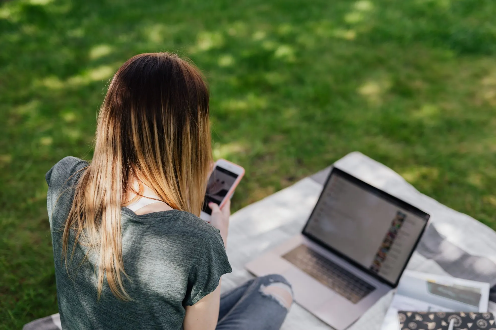Junge Person im Park, über die Schulter fotografiert, mit Smartphone und Laptop